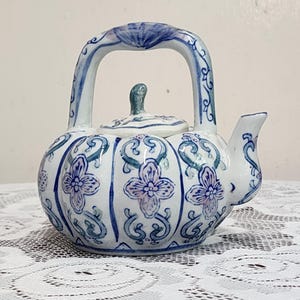 Puede incluir: Una tetera de cerámica con cuerpo redondeado y acanalado que presenta patrones florales en azul y morado claro sobre un fondo blanco. La tetera tiene un asa alta arqueada, una tapa pequeña con pomo y un pico corto, decorados con volutas azules.