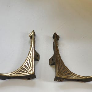 Op de afbeelding: Twee antieke messing plankdragers. Elke beugel heeft een decoratief zonnestraalontwerp en een gebogen, voetachtig basis. De beugels hebben een warme, verouderde messingkleur, ideaal voor vintage of retro decor.