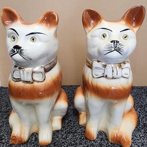 May include: Two ceramic cat figurines. The cats are white with brown ears, markings, and legs. They have black whiskers, green eyes, and a bow tie. The figurines are sitting upright.