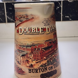Può includere: Una brocca in ceramica con un'illustrazione colorata di un complesso di birrifici. Il testo recita DOUBLE DIAMOND, TRADE MARK, Since 1876 e BURTON ON TRENT, IND COOPE & CO LTD. Il design include edifici in mattoni rossi e strutture industriali.