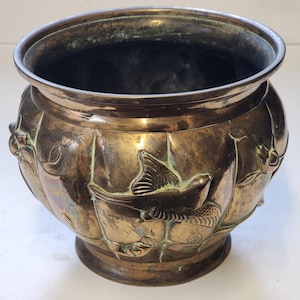 May include: An antique bronze planter with a wide rim and a rounded body. The planter features embossed bird and leaf designs around its exterior. The interior is dark, and the planter sits on a small, circular base.