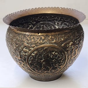 Op de afbeelding: Een sierlijke, bronskleurige metalen kom met ingewikkelde bloemen- en figuurreliëfs. De kom heeft een brede, geschulpte rand en een afgeronde body. Het gedetailleerde ontwerp suggereert een decoratief of ceremonieel doel.