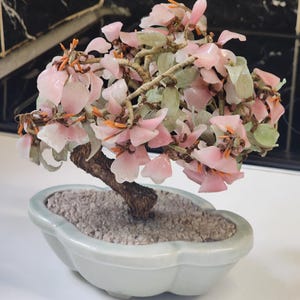 May include: A decorative bonsai tree sculpture featuring light pink stone flower petals, pale green stone leaves, and thin orange stamens. The tree has a textured brown trunk and is set in a light green, scalloped ceramic pot filled with small gray stones.