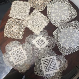 May include: A collection of nine off-white lace doilies featuring various intricate needlework patterns, including floral motifs, geometric openwork, and scalloped edges, arranged in a group with square and circular shapes of differing sizes.