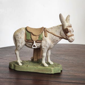 Puede incluir: Figura de cerámica de un burro con montura. El burro es blanco con manchas marrones y una montura marrón con ribete verde. Se encuentra sobre una base verde con un tronco de árbol. La figura está sobre una superficie de madera oscura.