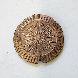 Può includere: Un posacenere rotondo color bronzo con un motivo centrale a forma di raggio di sole. Il posacenere presenta una serie di forme a forma di foglia radianti e un bordo esterno circolare con scanalature verticali. Una piccola tacca è visibile sul bordo.