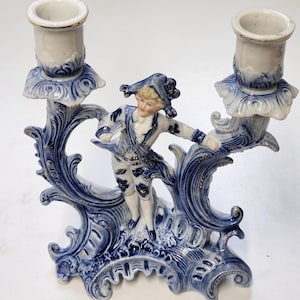 Puede incluir: Un candelabro doble de porcelana azul y blanca con una figura de un hombre con un disfraz. El candelabro tiene dos soportes con detalles florales y ribetes dorados. La base es ornamentada con patrones arremolinados.