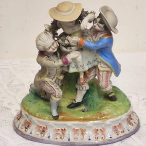 Könnte beinhalten: Porzellanfigur, die drei Figuren in Kleidung aus dem 18. Jahrhundert darstellt. Die zentrale Figur trägt ein geblümtes Kleid und einen Strohhut, während zwei Figuren sie in blauen und beigen Mänteln flankieren. Der Sockel ist mit floralen Mustern verziert.