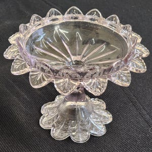 Op de afbeelding: Een heldere, lavendelkleurige glazen schaal op voet. De schaal heeft een geschulpte rand en een gedetailleerde, bloemachtige basis. De kom heeft een stervormig patroon dat vanuit het midden uitstraalt.