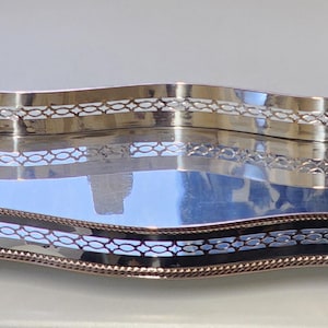 May include: A long, silver serving tray with a wavy design. The tray features a decorative openwork border and a reflective surface. The edges are trimmed with a beaded design, and the tray rests on small feet.