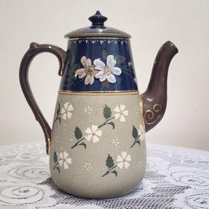 Może przedstawiać: Ceramiczny imbryk z ciemnoniebieską górną częścią z białymi akcentami kwiatowymi oraz teksturowanym jasnoszarym korpusem ozdobionym białymi kwiatami i zielonymi liśćmi. Imbryk posiada ciemnobrązowy uchwyt, pasujący dzióbek oraz pokrywkę.