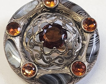 May include: A round, silver-toned brooch with a central, large, brown gemstone surrounded by smaller, orange gemstones. The brooch features a decorative, openwork design and is accented with oval-shaped, black and white striped stones.