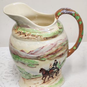 Può includere: Brocca in ceramica color crema con un paesaggio. La brocca ha un manico marrone con dettagli di foglie verdi. Il corpo mostra una scena di montagna, una carrozza trainata da cavalli e il testo "THE IRISH JAUNTING CAR".