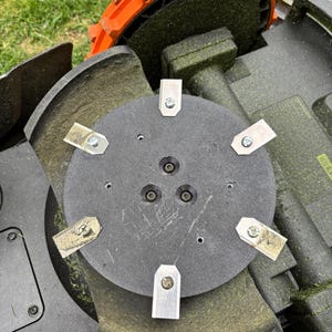 May include: A circular black mower blade disc featuring six small, rectangular metal cutting blades attached with screws. The disc is mounted on a dark gray robotic lawn mower chassis with a large orange wheel visible at the top edge.