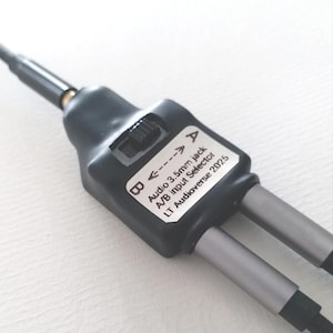 Peut inclure: Un sélecteur d'entrée audio jack 3,5 mm noir A/B avec deux câbles de sortie argentés. L'appareil comporte un interrupteur et une étiquette indiquant "Audio 3.5mm jack A/B input Selector LT Audioverse 2025".