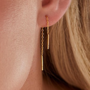 May include: Gold-colored threader earrings. Each earring features a delicate chain that passes through the ear and suspends a slender, vertical bar. The earrings are shown against a green background.