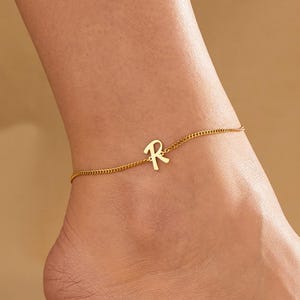 May include: A gold-colored anklet with the letter "R" charm. The delicate chain is draped around the ankle. The anklet is displayed on a light brown background.
