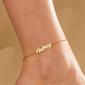 May include: A gold-colored anklet with the name "Aubrey" in a cursive font. The anklet is worn on an ankle, with the foot flexed. The background is a neutral, light tan color.