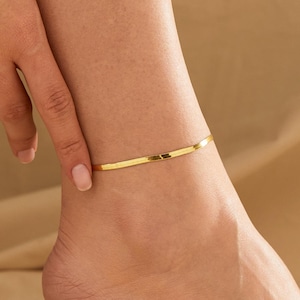 May include: A gold-colored, flat herringbone chain anklet. The anklet is worn around the ankle. The image is a close-up shot of the ankle and foot. The background is a neutral, light tan color.