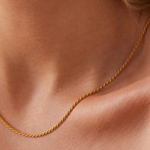 May include: A delicate gold-colored rope chain necklace. The necklace is displayed on a person's neck and chest. The chain has a twisted rope design and is a simple, elegant piece of jewelry.