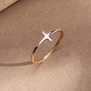 May include: A gold ring with a cross design. The ring has a thin band and a simple cross shape. The cross is silver, contrasting with the gold band. The ring is displayed on a neutral-toned surface.