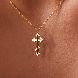 May include: A gold-toned necklace featuring a delicate chain and a small, intricate Eastern Orthodox cross pendant. The cross has a distinct three-bar design with rounded ends, suspended from a simple link chain that rests against the collarbone.