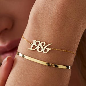 May include: Two gold bracelets are displayed on a wrist. The top bracelet features the number "1986" in a delicate script. The lower bracelet is a simple, textured band. The bracelets are set against a soft, neutral background.