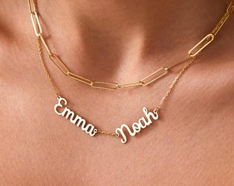 May include: A gold-colored, layered necklace with the names "Emma" and "Noah" in cursive script. The top layer features a chain with elongated rectangular links. The necklace is displayed on a person's chest.