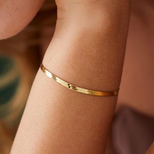 May include: A gold-colored bracelet with a small, dark accent is worn on a person's arm. The bracelet is thin and appears to be made of metal. The skin tone is light, and the focus is on the jewelry.