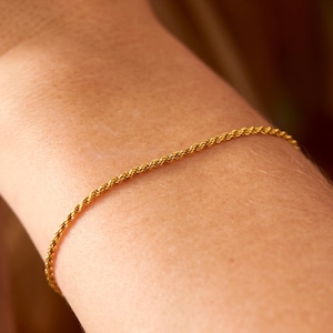 May include: A delicate gold-colored rope chain bracelet. The bracelet is worn on a wrist, and the chain has a twisted design. The bracelet is a simple, elegant piece of jewelry.