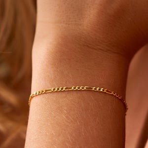 May include: A delicate gold-colored bracelet with a Figaro chain design. The bracelet is worn on a wrist, showcasing its fine links and subtle shine. The jewelry piece is a simple yet elegant accessory.