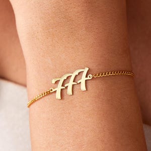 May include: A gold-colored bracelet with the number "777" in a cursive font. The bracelet is attached to a delicate chain. The bracelet is worn on a person's wrist.