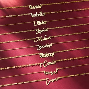 May include: Gold-colored name necklaces with various names, including Scarlett, Isabella, Olivia, Sophia, Madison, Brooklyn, Aubrey, Camila, Hazel, and Emma. The necklaces are displayed on a burgundy background.