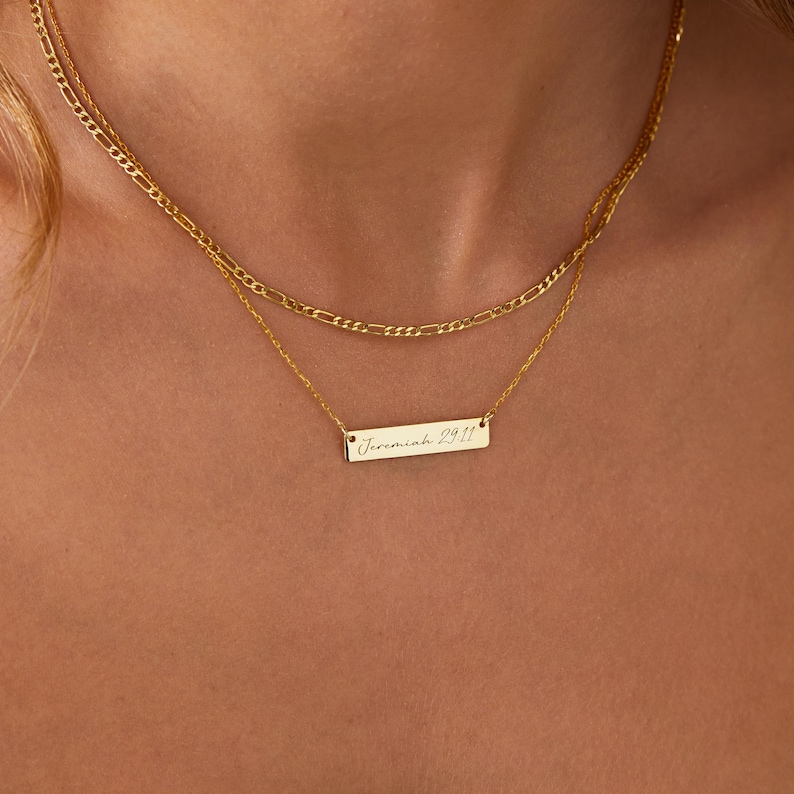 May include: A layered gold necklace set. The top chain is a curb chain, and the bottom chain features a rectangular bar pendant. The pendant is engraved with the text "Jeremiah 29:11".