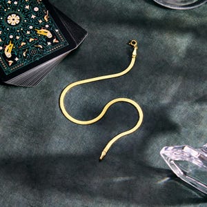 K&ouml;nnte beinhalten: Eine goldfarbene Fischgr&auml;ten-Kettenhalskette mit Verschluss, die auf einer dunkelgr&uuml;nen Oberfl&auml;che liegt. Ein Tarotkarten-Deck mit dekorativem Design befindet sich im Hintergrund. Eine klare Glasschale ist ebenfalls sichtbar.