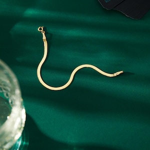 K&ouml;nnte beinhalten: Ein goldfarbenes Fischgr&auml;ten-Armband mit Karabinerverschluss, pr&auml;sentiert auf einer dunkelgr&uuml;nen Oberfl&auml;che. Das Armband hat ein flaches, schlangenartiges Design. Der Verschluss ist klein und rund.