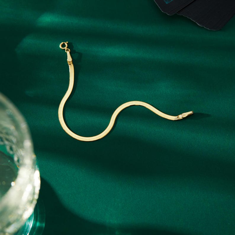 K&ouml;nnte beinhalten: Ein goldfarbenes Fischgr&auml;ten-Armband mit Karabinerverschluss, pr&auml;sentiert auf einer dunkelgr&uuml;nen Oberfl&auml;che. Das Armband hat ein flaches, schlangenartiges Design. Der Verschluss ist klein und rund.