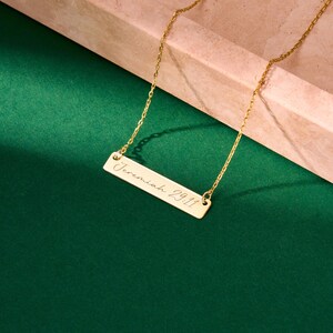 May include: A gold-colored bar necklace with the inscription "Jeremiah 29:11" in a delicate script. The rectangular pendant hangs from a fine chain, set against a green background.