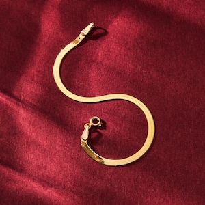 K&ouml;nnte beinhalten: Ein goldfarbenes Fischgr&auml;tenarmband mit kleinem Verschluss. Das Armband liegt auf einer tiefroten, strukturierten Oberfl&auml;che. Am Ende des Armbands befindet sich ein kleiner, klarer Stein.