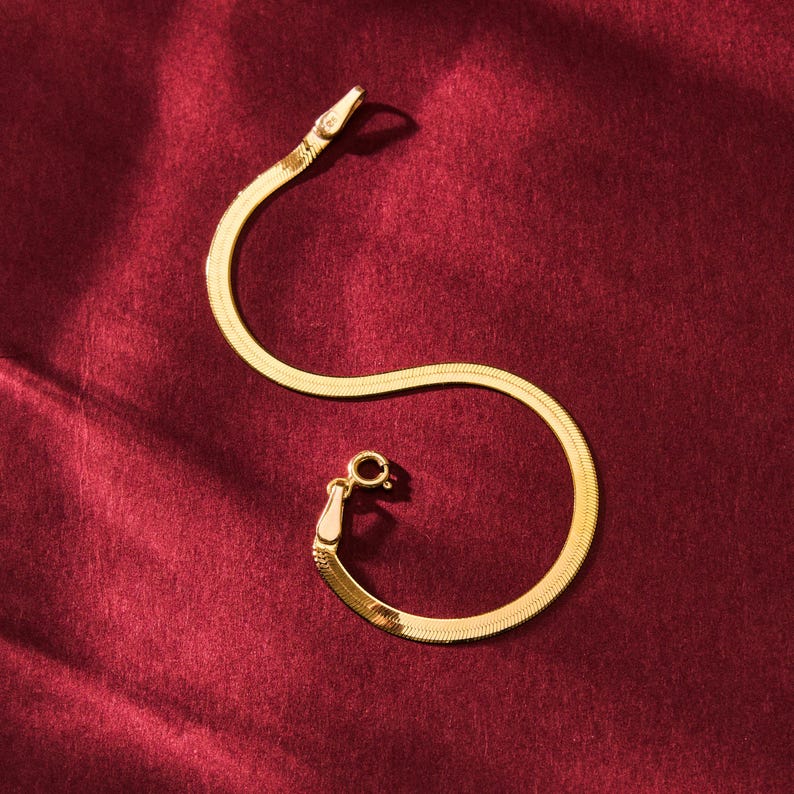 K&ouml;nnte beinhalten: Ein goldfarbenes Fischgr&auml;tenarmband mit kleinem Verschluss. Das Armband liegt auf einer tiefroten, strukturierten Oberfl&auml;che. Am Ende des Armbands befindet sich ein kleiner, klarer Stein.