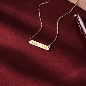 May include: A gold-colored necklace with a rectangular pendant. The pendant is inscribed with the text "John 3:16". The necklace is displayed on a burgundy surface, with a pen visible in the background.