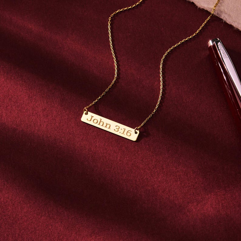 May include: A gold-colored necklace with a rectangular pendant. The pendant is inscribed with the text "John 3:16". The necklace is displayed on a burgundy surface, with a pen visible in the background.