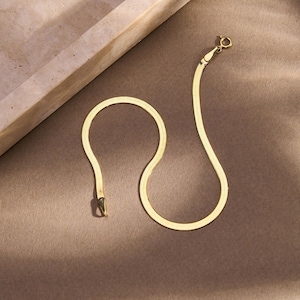 K&ouml;nnte beinhalten: Ein goldfarbenes Fischgr&auml;ten-Armband mit Karabinerverschluss, das auf einer braunen Oberfl&auml;che liegt. Das Armband besteht aus kleinen, flachen, ineinandergreifenden Gliedern, die ein glattes, flexibles Design ergeben. Der Verschluss ist am Ende der Kette sichtbar.