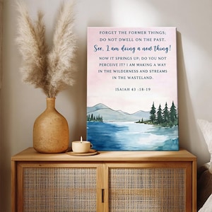 以下が含まれることがあります： イザヤ書43:18-19からのテキストが書かれた、湖と山の水彩画の風景が描かれた額入りのキャンバスプリント。プリントは、乾燥したパンパスグラスの花瓶とろうそくが灯された木製のキャビネットに飾られています。