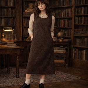 May include: A brown, knee-length dress with a textured weave, featuring a scoop neckline and pockets. The dress is worn over a light-colored collared shirt. The person is wearing black shoes and white socks. The background includes bookshelves and a wooden table.