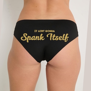 May include: Black underwear with the text "IT AINT GONNA Spank Itself" in gold glitter lettering. The underwear is a brief style, and the image shows the back of the garment.