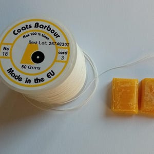 Pode incluir: Um carretel de linha de linho branco Coats Barbour, com o texto "Flax 100% Linen" e "Made in the EU". Dois pequenos blocos retangulares amarelos estão ao lado da linha. O carretel está rotulado como "50 Grms".