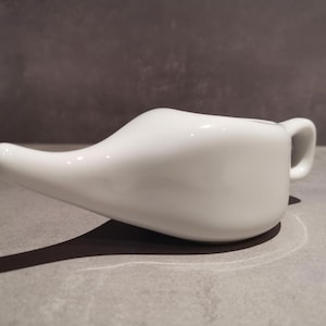 Könnte beinhalten: Eine weiße Keramik-Neti-Kanne mit langem Ausguss und kleinem Griff. Die glatte, glänzende Oberfläche reflektiert das Licht, und die Kanne wirft einen Schatten auf die graue Oberfläche darunter. Dieses Nasenspülgerät wird zur Sinuspflege verwendet.