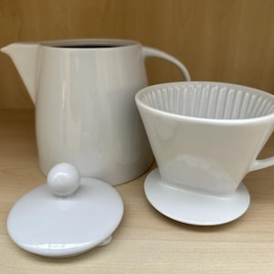 Könnte beinhalten: Ein weißes Keramik-Pour-Over-Kaffee-Set, bestehend aus einer Karaffe, einem Filterhalter und einem Deckel mit einem kleinen Knopf. Das Set wird auf einer hellen Holzoberfläche präsentiert. Die Karaffe hat einen Griff und einen Ausguss.