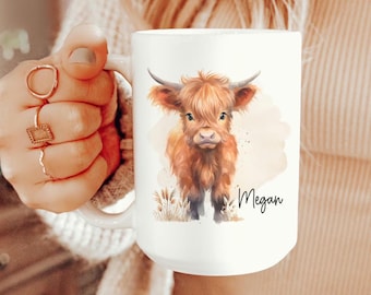 May include: White ceramic mug featuring a watercolor illustration of a Highland cow with brown fur and horns. The mug has the name "Megan" written in script. The mug is held by a person wearing a beige sweater and blue jeans.